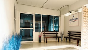 Zona de recepción de un hotel con puerta de vidrio, dos bancos de madera, un tapete de bienvenida y un letrero con el nombre del hotel.