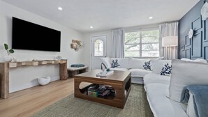 Un soggiorno moderno con TV a schermo piatto, un tavolino in legno contenitore, un divano bianco con cuscini decorati e un'ampia finestra con tende.