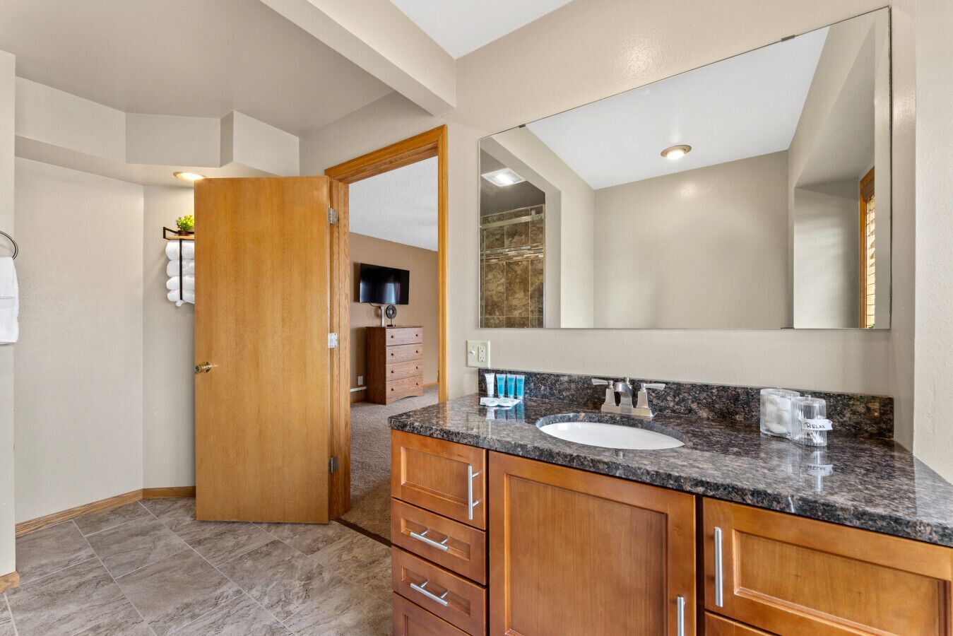 A bathroom with a granite countertop, a sink, a large mirror, wooden cabinets, and a door leading to another room.