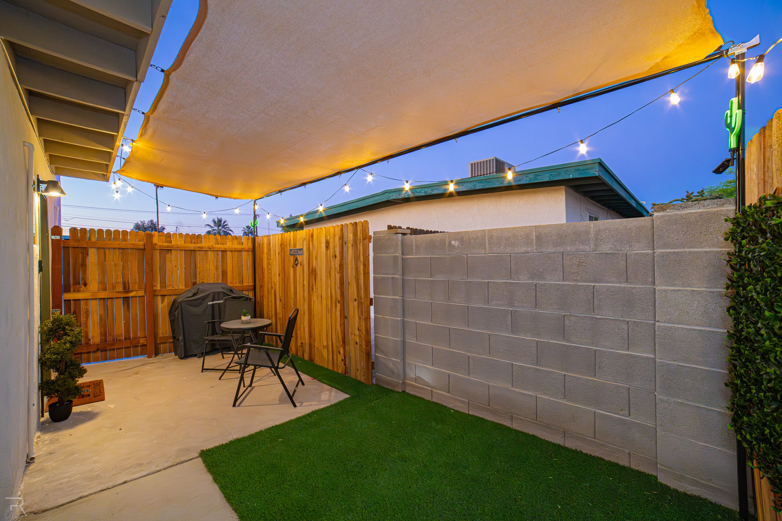 Un patio trasero con zona de barbacoa, césped artificial, valla de madera y luces colgantes.