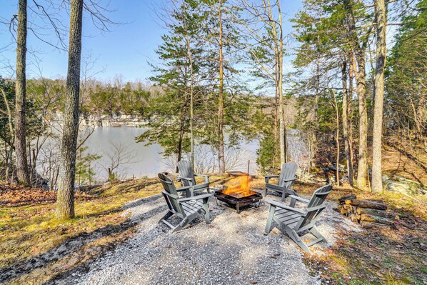 A campfire with four Adirondack chairs arranged around it, set on a gravel area with trees and a lake in the background. - Cumberland Lake Retreat: Furnished Deck & Grill! (Burnside)