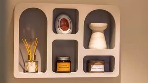 A wall-mounted shelf with four compartments containing a white vase, a white candle holder, a jar of apple cinnamon, and a container of reed diffusers. - Mabulle Originality & Comfort in the Dijon's Heart (Dijon)
