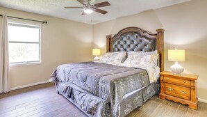Une chambre à coucher avec une tête de lit capitonnée, un lit recouvert d’une couvre-lit à motifs foncés, deux tables de chevet avec des lampes, un ventilateur de plafond et une fenêtre avec des rideaux.