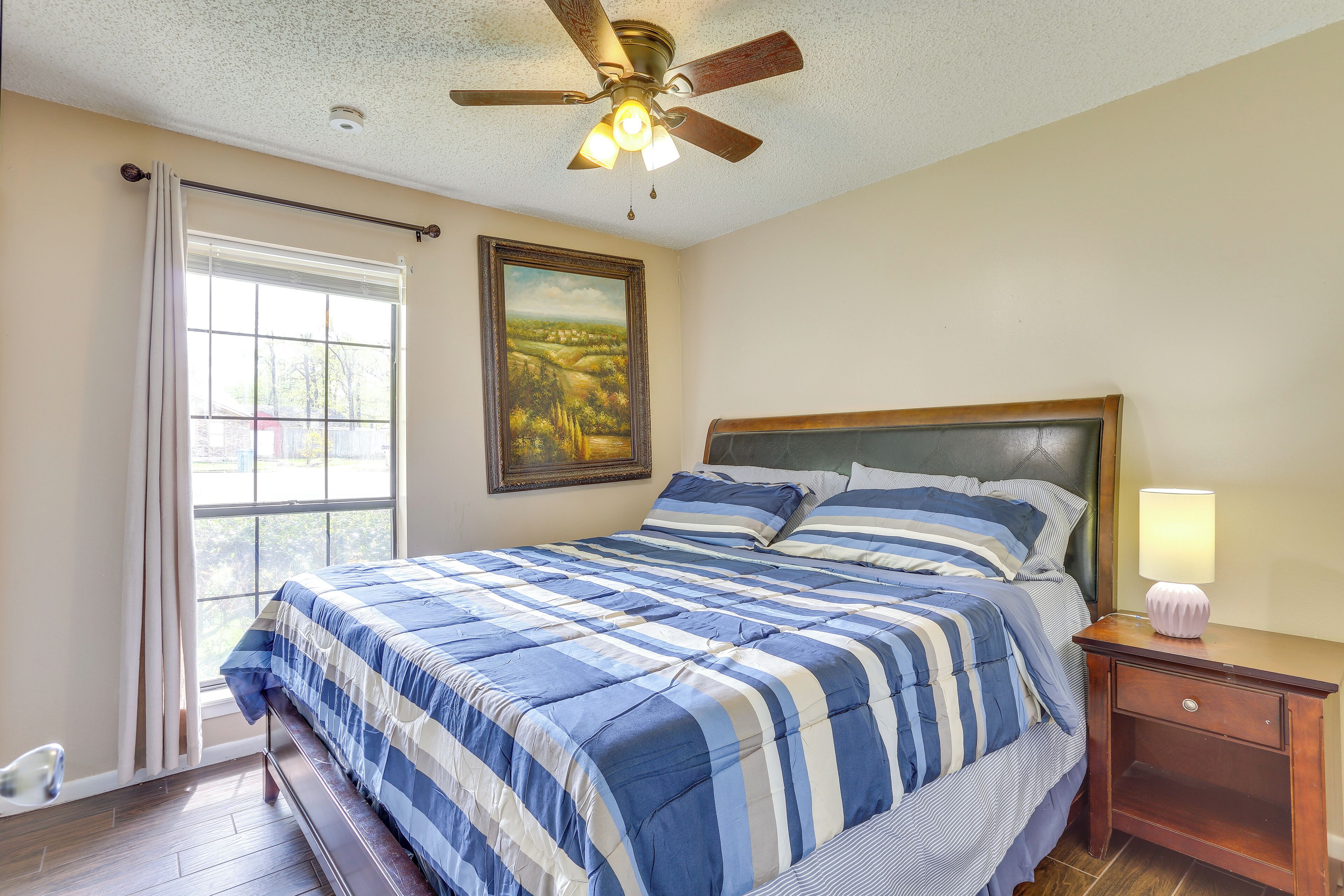 A bedroom with a bed, a nightstand, a ceiling fan, a window with curtains, and a landscape painting on the wall.