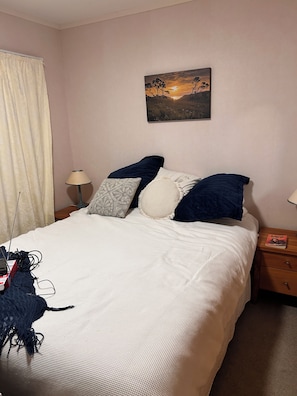 A neatly made bed with a white comforter and multiple pillows, a painting of a sunset on the wall, a lamp on a bedside table, and a book on a nightstand. - Peaceful & restorative Awhitu Peninsula retreat-80 minutes from Auckland CBD (Awhitu)