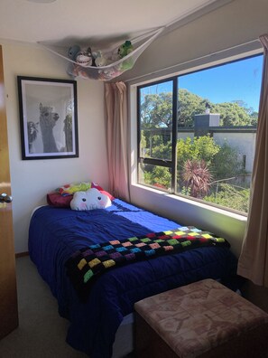A bedroom with a large window, a bed with a blue bedspread, a colorful quilt, and a framed picture on the wall.
