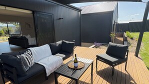 An outdoor patio with a black sofa, a wooden table, and a wine bottle. - Goolwa Beach Pavilion (Goolwa Beach)