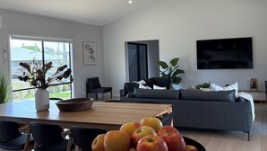 A modern living room with a dining area, featuring a large black bowl of apples on the table, a grey sofa, and a flat-screen TV mounted on the wall. - Goolwa Beach Pavilion (Goolwa Beach)