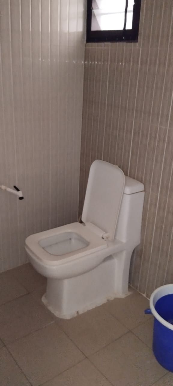 A small, compact bathroom with a white toilet, a blue bucket, and a window above the toilet.