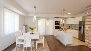A modern kitchen with white cabinetry, a central island, and a dining area with a white table and chairs. - Villa Kate - Dubrovnik by Villas Guide (Dubrovnik)