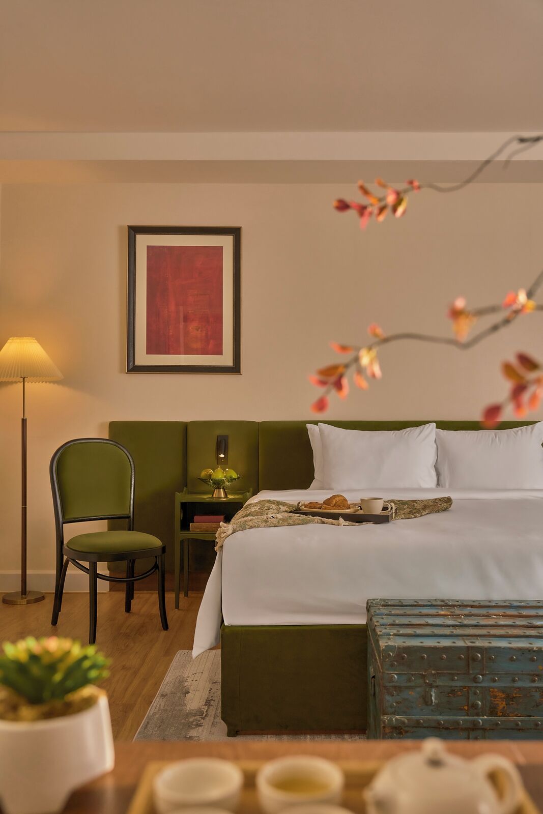 A hotel room with a bed, a chair, a bedside table with a lamp, a framed picture on the wall, and a small potted plant.