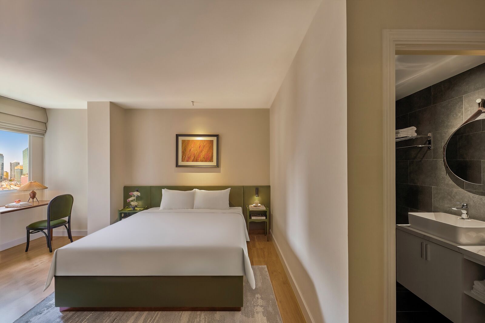 Ein modernes Hotelzimmer mit einem großen Bett, einem Schreibtisch mit Stuhl, Blick auf die Stadt und einem Badezimmer mit Badewanne und Waschbecken.