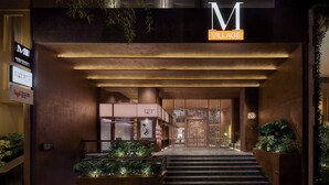 Una entrada de hotel moderna con un llamativo letrero 'M Village', una escalera que conduce al vestíbulo y un letrero que dice 'Cada Medio Año' indicando un programa de fidelización.