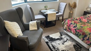 A hotel room with a gray sofa, a small table with a plant, a gray armchair, a bed with a floral bedspread, and a window with a view of buildings.