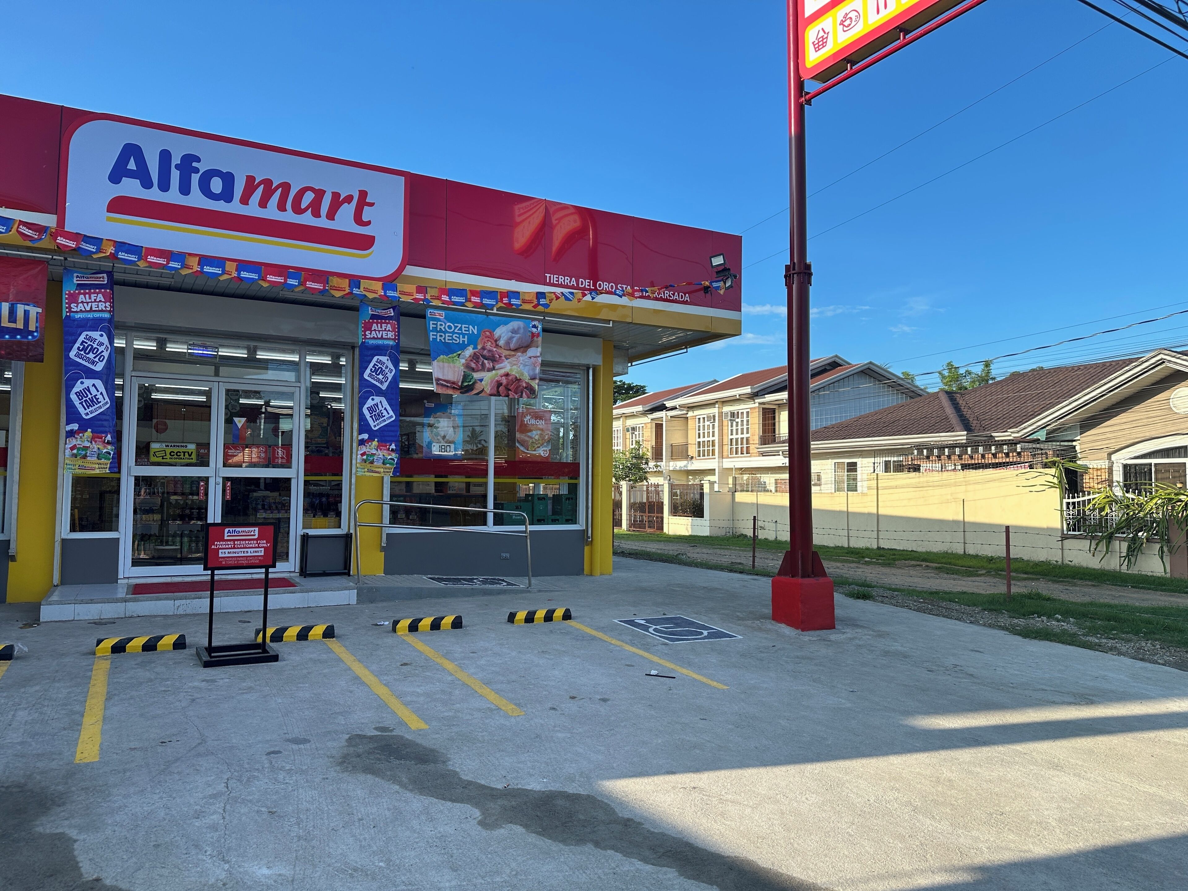 Fassade eines Alfamart-Geschäfts mit freiem Eingang, Werbeplakaten und einem Parkplatz mit ausgewiesenen Stellplätzen.