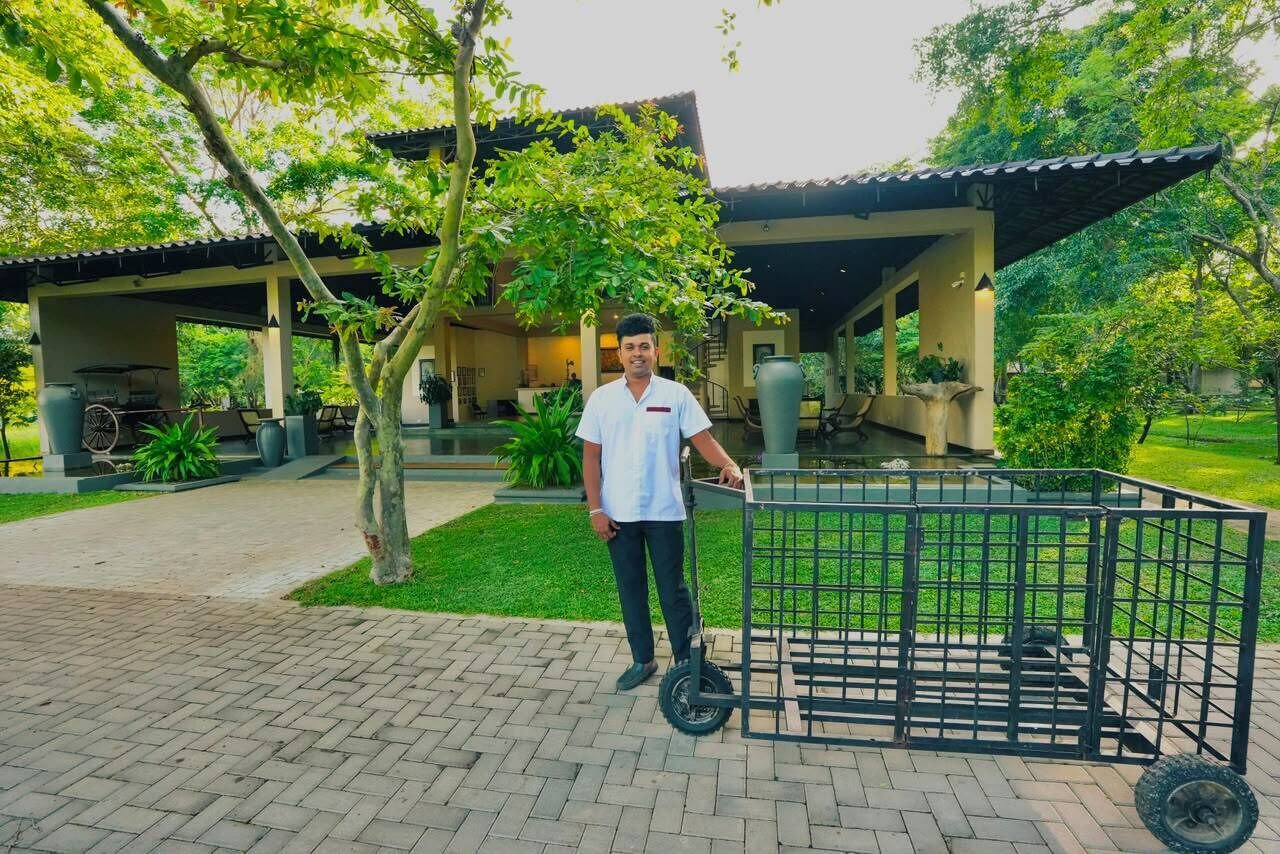 A man standing next to a black metal cart in a courtyard with a covered structure and greenery in the background.