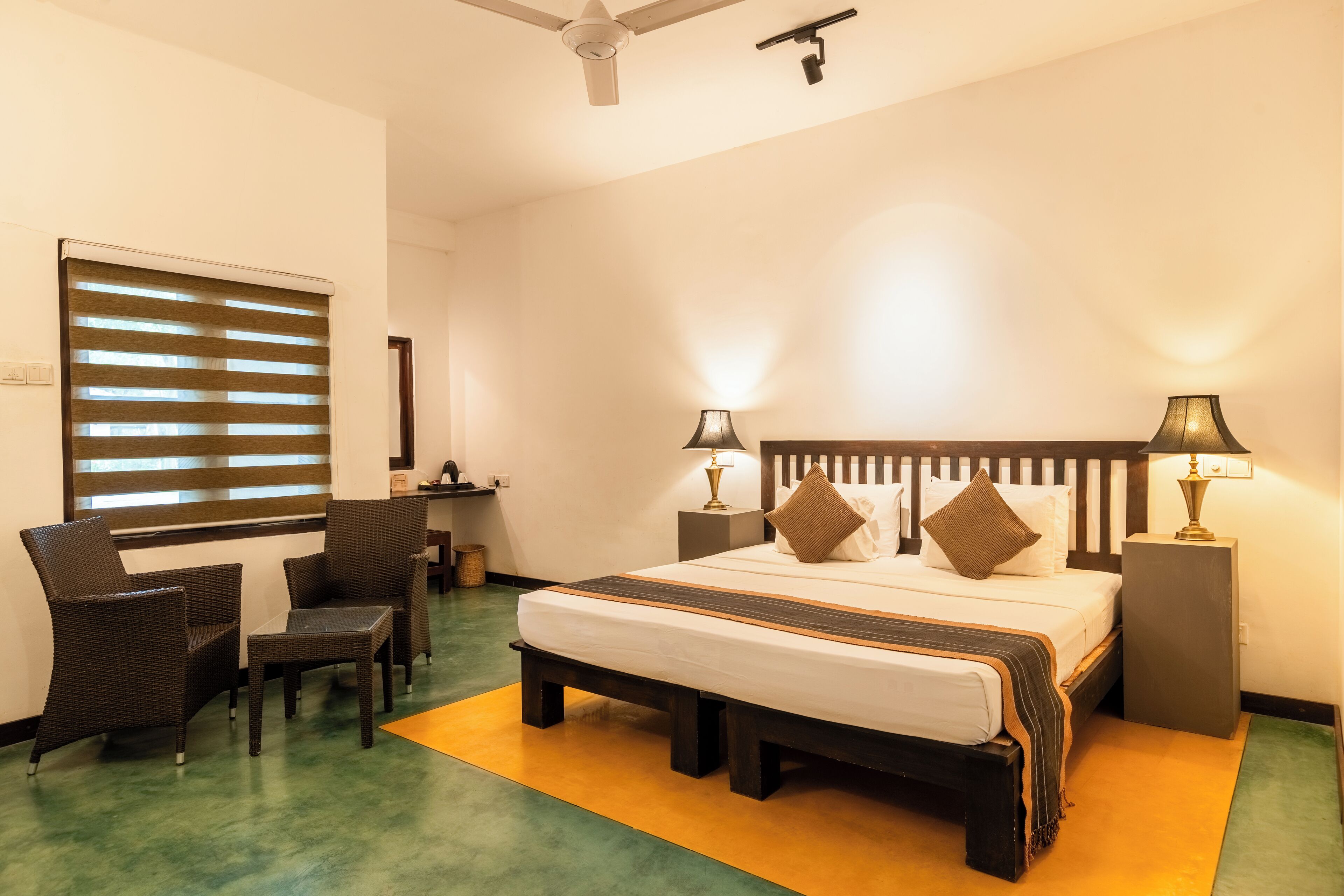 A hotel room with a bed, two bedside tables with lamps, a chair, a desk, and a window with blinds.