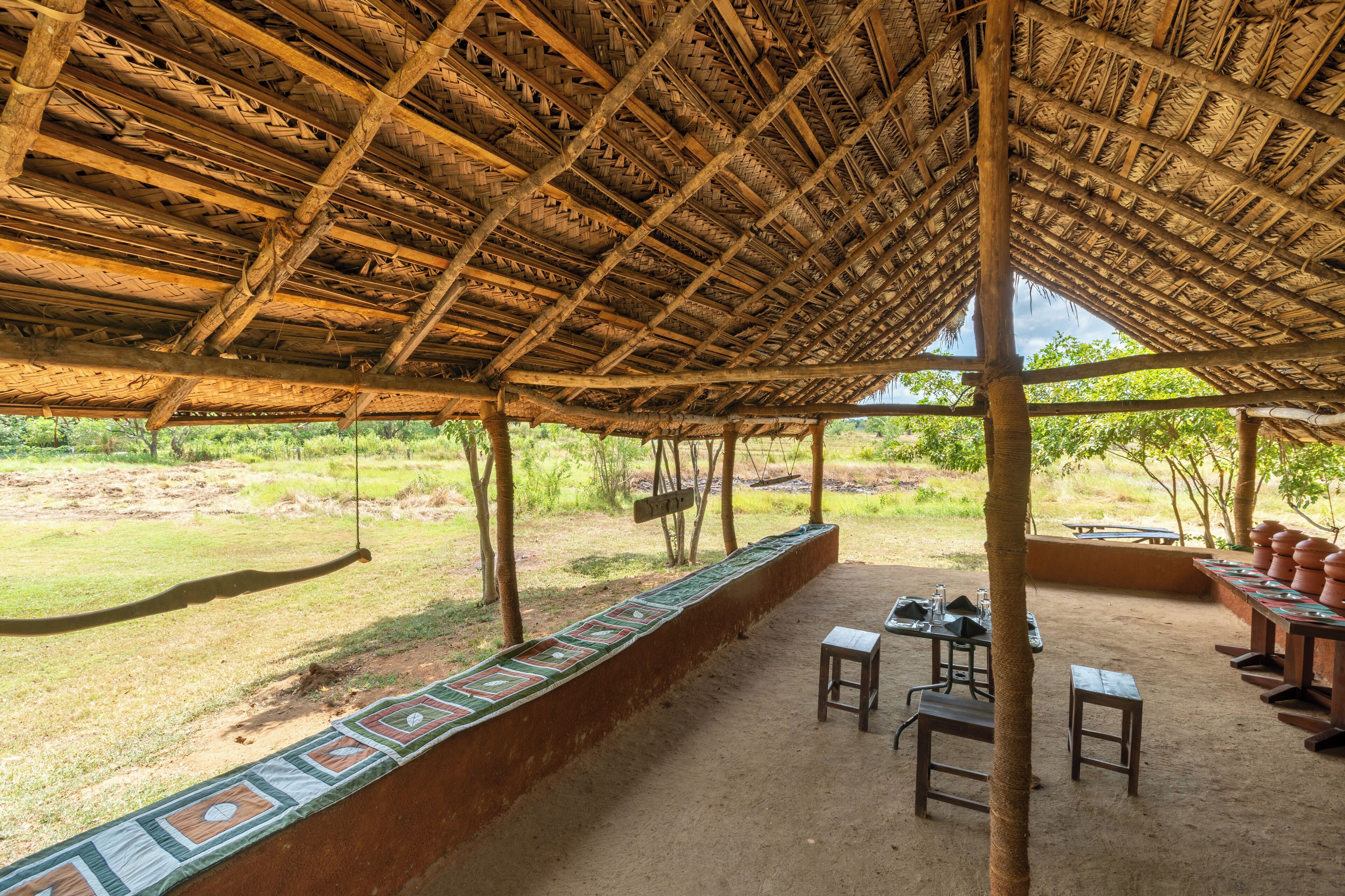 An outdoor covered area with a thatched roof, a long bench with a colorful edge, and a table with chairs.