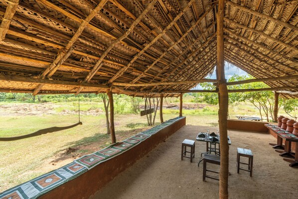 An outdoor covered area with a thatched roof, a long bench with a colorful edge, and a table with chairs. - Kassapa Lion Rock (Sigiriya)