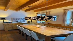 A dining area with a long wooden table, white chairs, and a hanging cow-patterned light fixture. - Country House Goldegg near Ski Slopes (Goldegg - Weng)