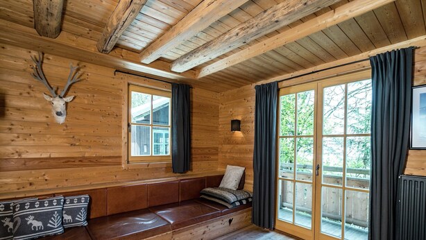 A log cabin interior with a wooden bench, a mounted deer head, and large windows.