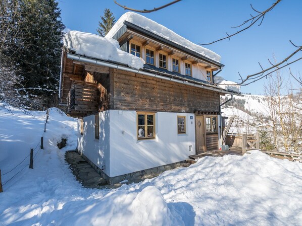 A two-story wooden chalet with a balcony, surrounded by snow.