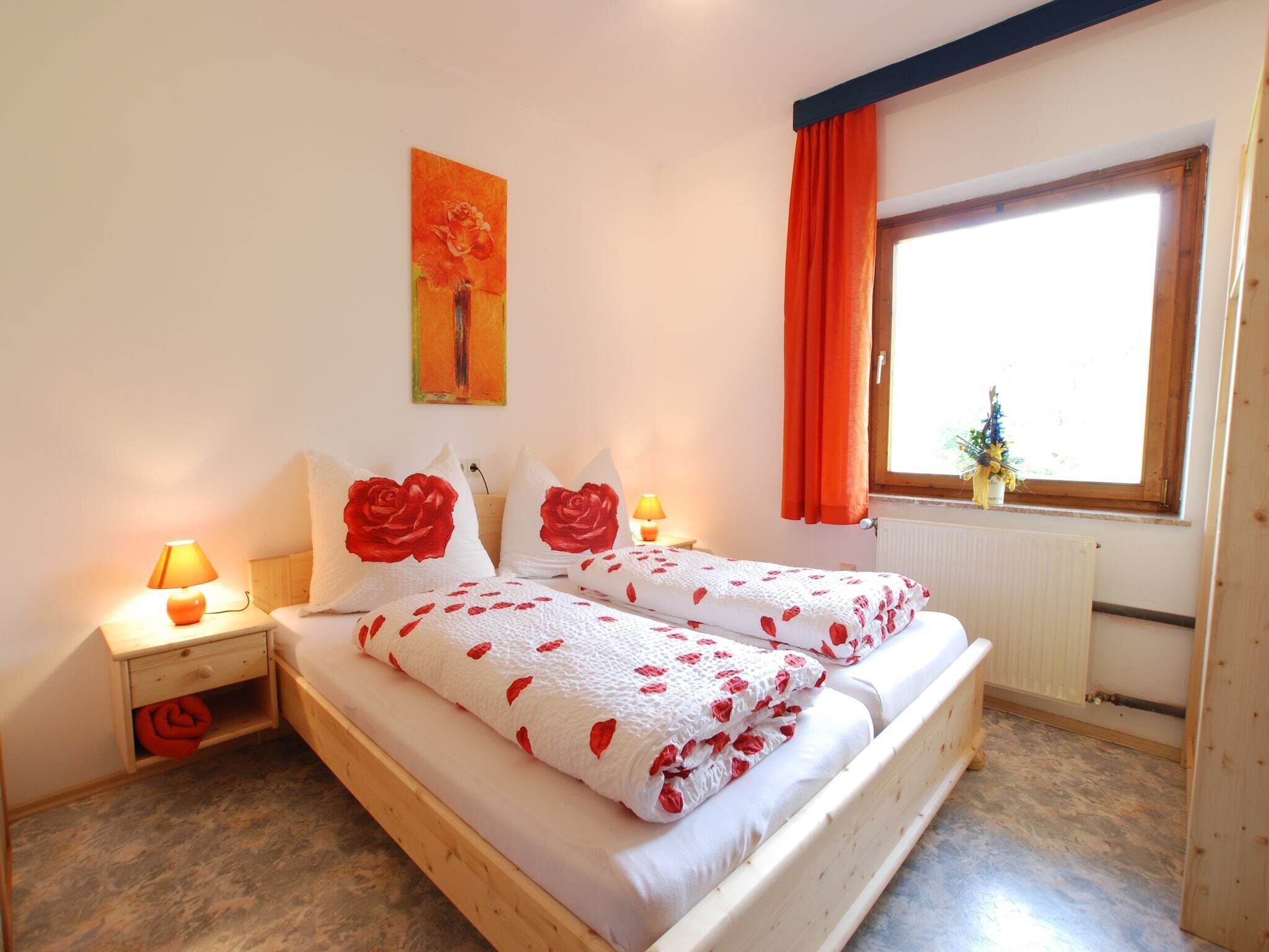 A bedroom with a wooden bed, two bedside tables with lamps, a window with orange curtains, and a painting on the wall.