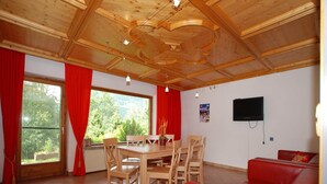 A dining area with a wooden table and chairs, a red sofa, a TV mounted on the wall, and a window with red curtains. - Chalet in Neukirchen near Ski Lifts (Neukirchen am Großvenediger)
