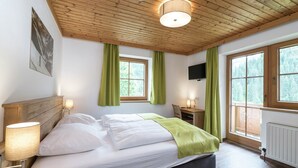 Quarto com teto de madeira, cama com roupa de cama branca, cortinas verdes, escrivaninha e televisão na parede.
