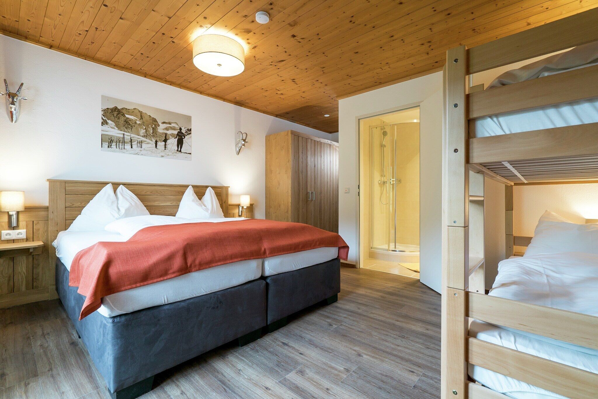 A bedroom with a wooden ceiling, a bed with white and red bedding, a wooden bunk bed, and a door leading to a bathroom.