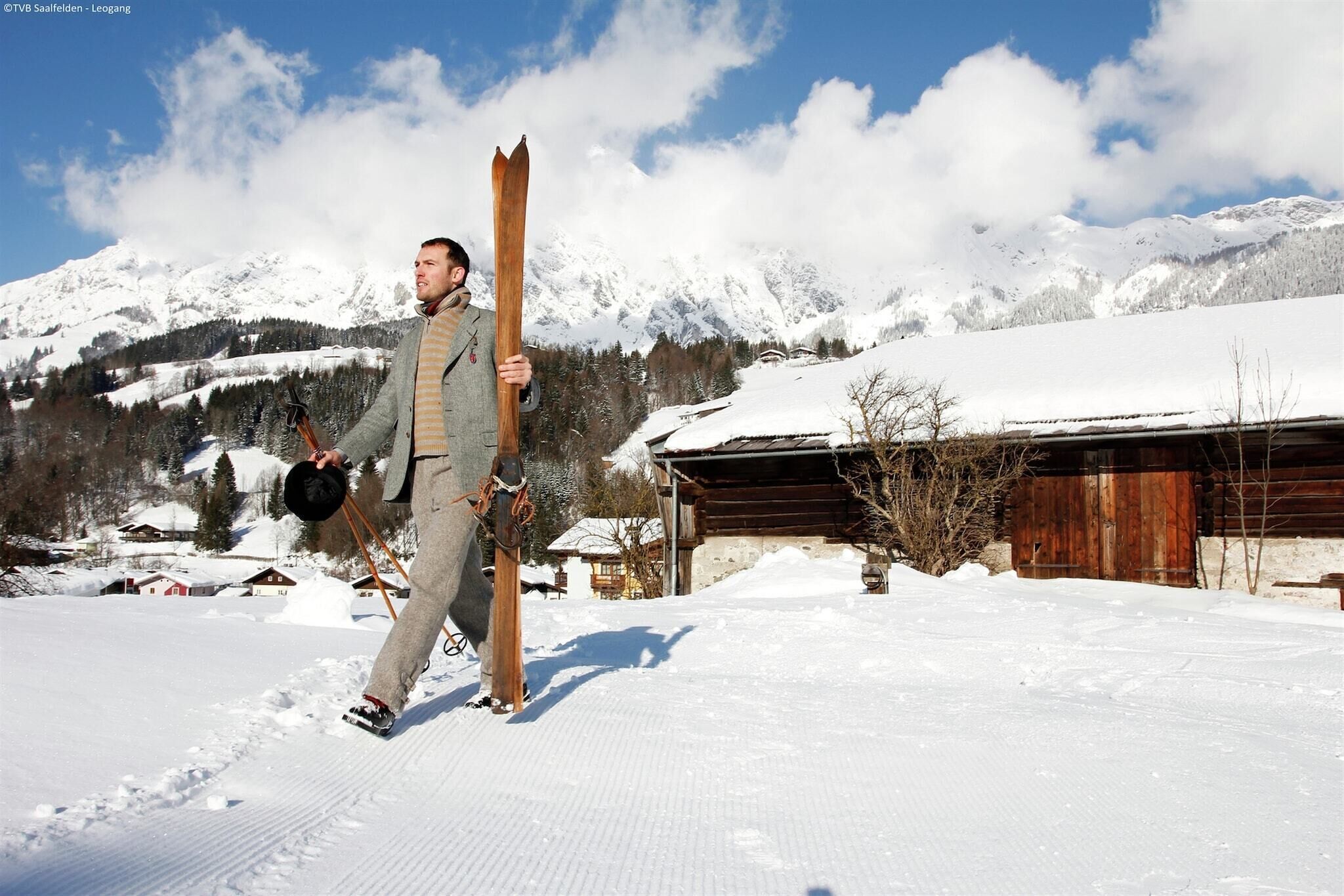 A person carrying skis and poles, walking on a snow-covered path with a wooden building and mountains in the background.