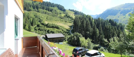 A balcony with a wooden floor, overlooking a valley with a road, a small building, and two cars parked on the grass.
