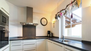 A modern kitchen with white cabinets, a stainless steel range hood, a built-in oven, and a window with decorative hanging items. - Holiday Home Brixen near Skiwelt Slopes (Brixen im Thale)