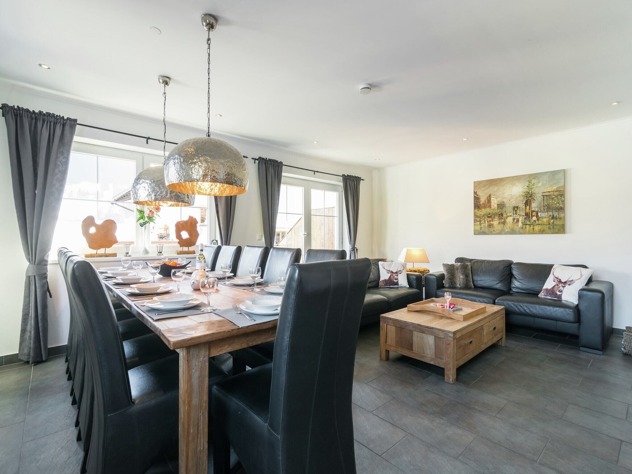 A spacious living area with a dining table, black leather chairs, a wooden coffee table, and a black leather sofa. There is a painting on the wall and a window with curtains.