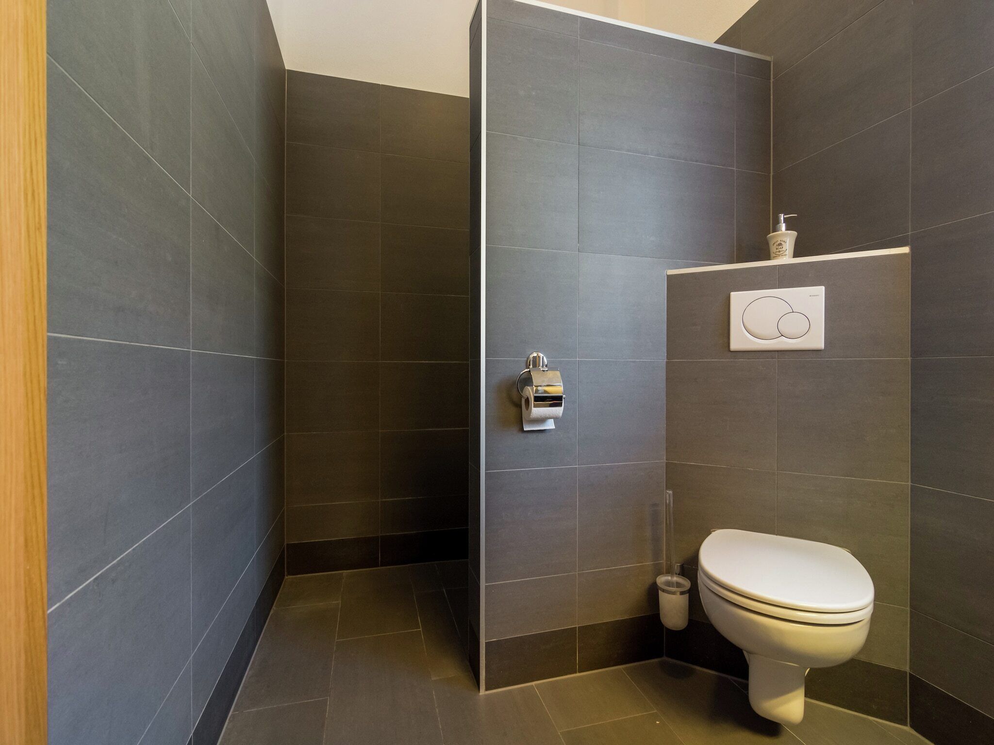 A modern bathroom with dark tiles, a toilet, a hand dryer, and a soap dispenser.