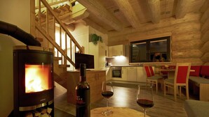 Un salon chaleureux avec un poêle à bois, une bouteille de vin et deux verres à vin posés sur une table en bois. Un escalier avec des rampes en bois mène à un espace cuisine équipé d’appareils modernes et doté d’une grande fenêtre.