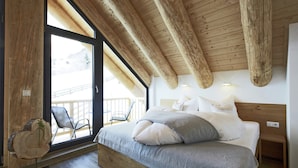 Une chambre avec un plafond en bois, une grande fenêtre, un lit avec des oreillers, une table de chevet et une chaise près de la fenêtre.