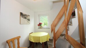 Un comedor con una mesa cubierta por un mantel amarillo, sillas de madera, un cuadro en la pared, una ventana con vistas a follaje y una escalera de madera.