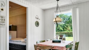 A cozy room with a bed, a dining table with chairs, a hanging light fixture, and a view of greenery outside.