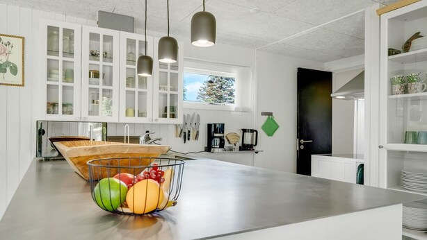 A modern kitchen with white cabinets, a central island with a fruit bowl, pendant lights, and a view of the outdoors.