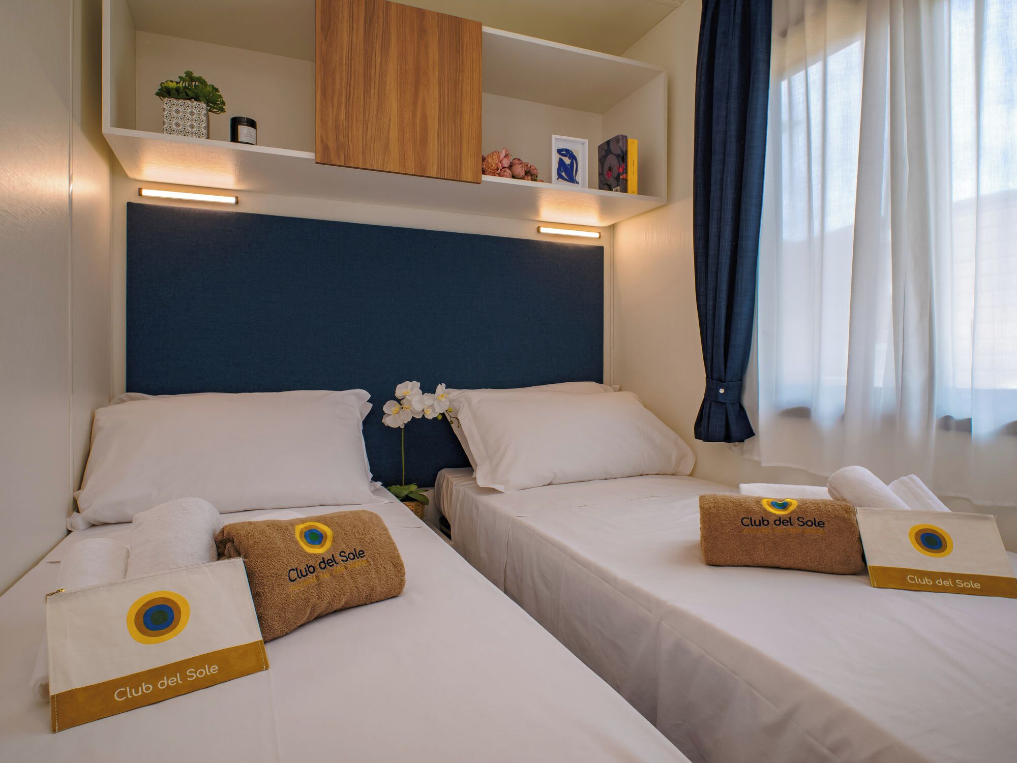 A hotel room with two beds, each with a white bedspread and pillows, and a blue headboard. There are two Club del Sole towels on each bed.