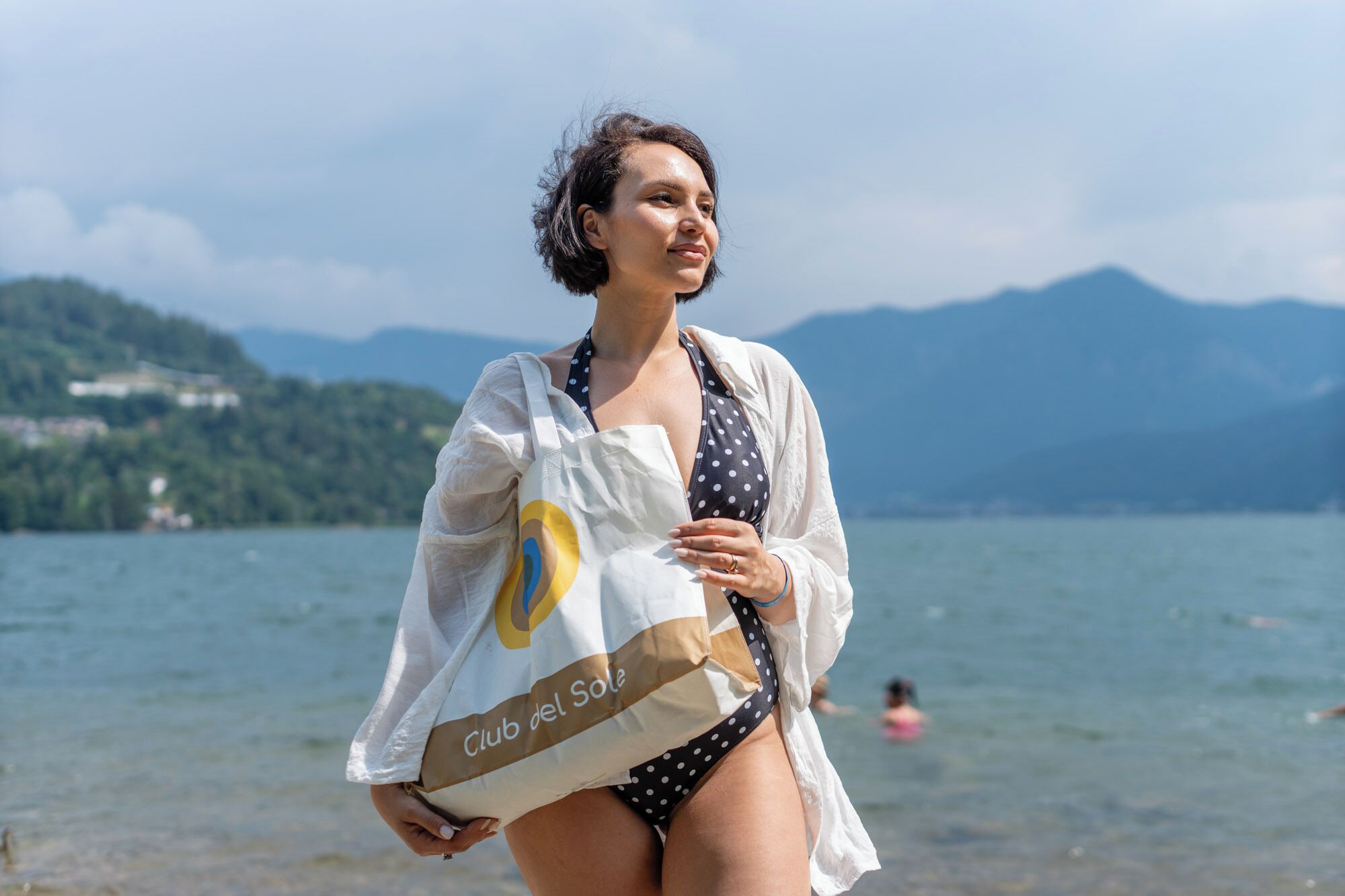 A person holding a bag with 'Club del Sole' text, standing by a lake with mountains in the background.