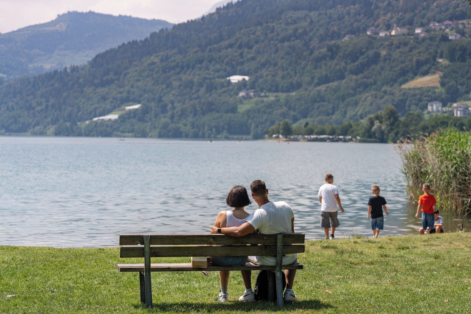 A lake with a bench, people walking, and a hilly landscape in the background.