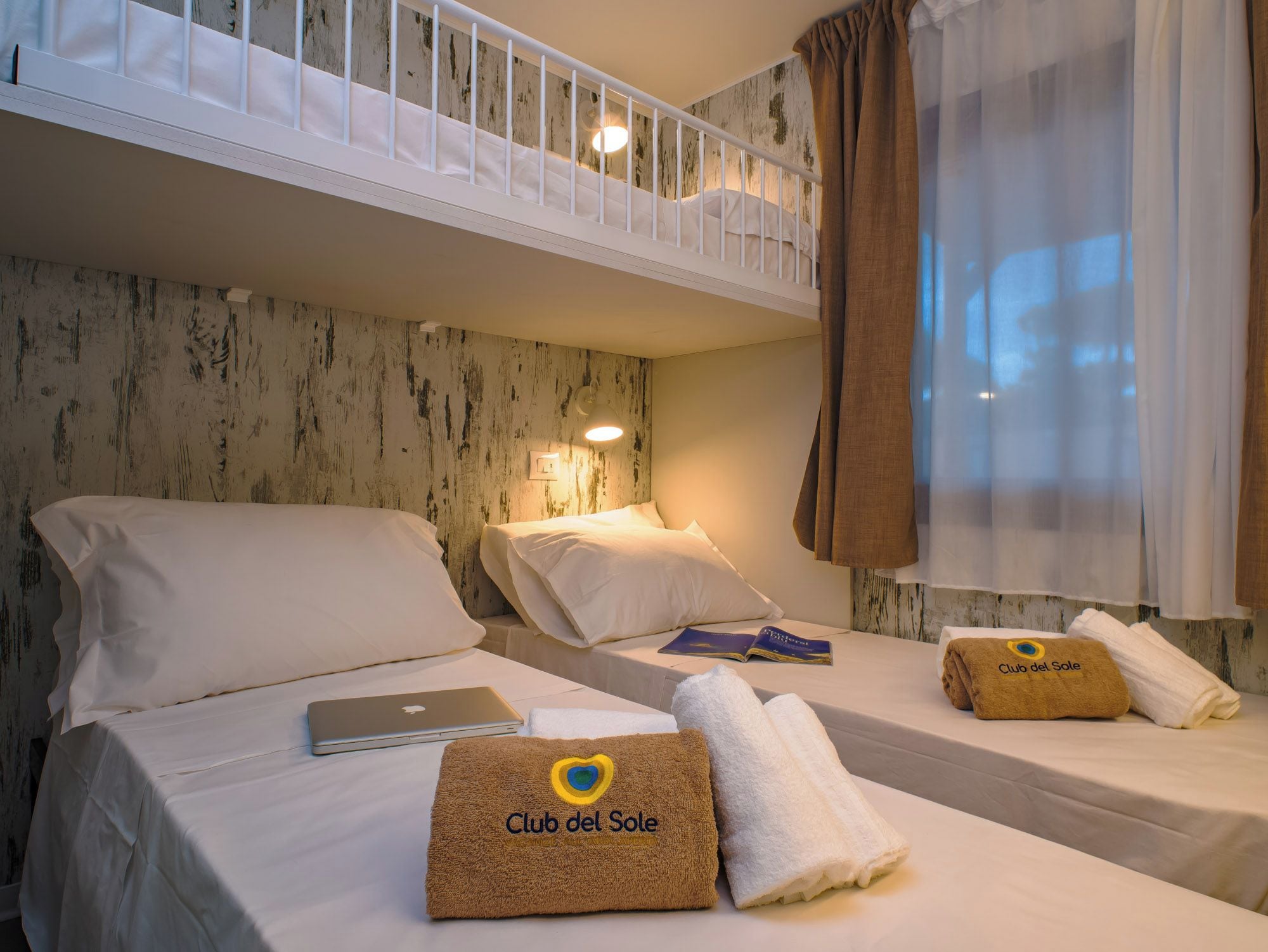 Ein Hotelzimmer mit Etagenbetten, einem Laptop, Handtüchern mit 'Club del Sole'-Logo und einem Vorhangfenster.