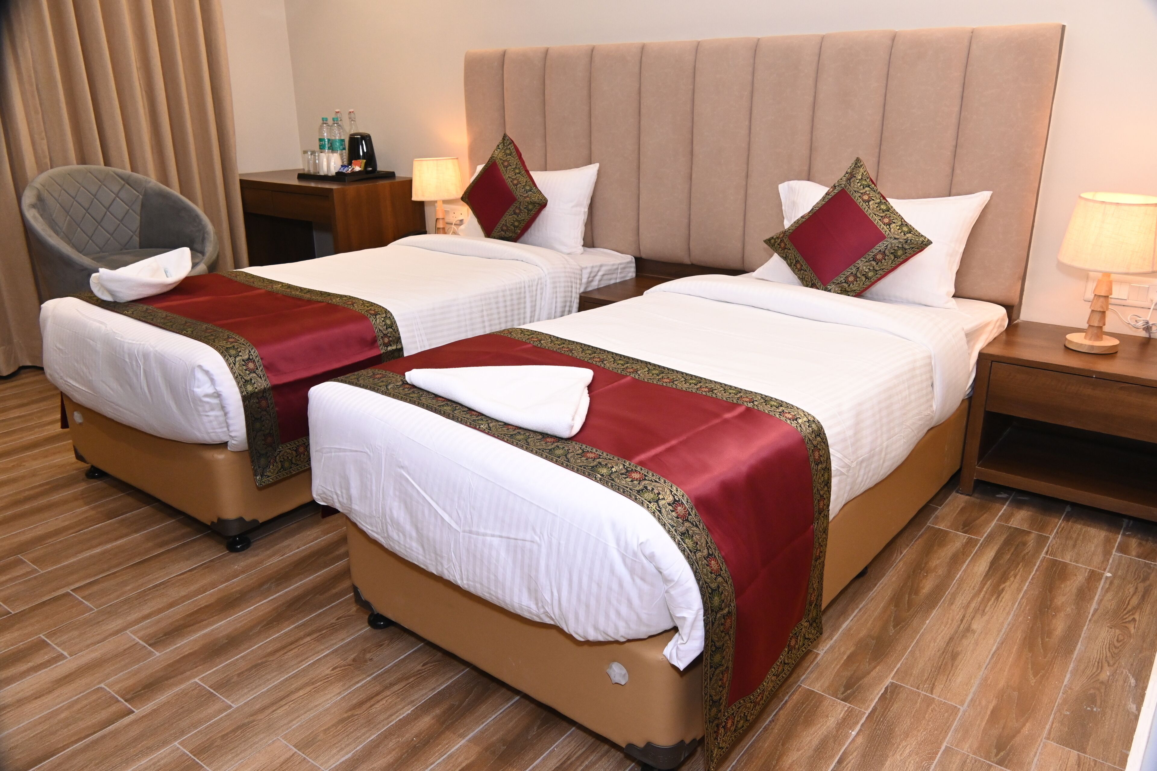 A hotel room with two beds, a chair, a nightstand with a lamp, and a window with curtains.