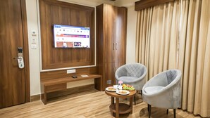 A modern room with a wooden wardrobe, a mounted TV, a round wooden table with a fruit bowl, and two grey armchairs. - Hotel The Rising Banaras (Varanasi)