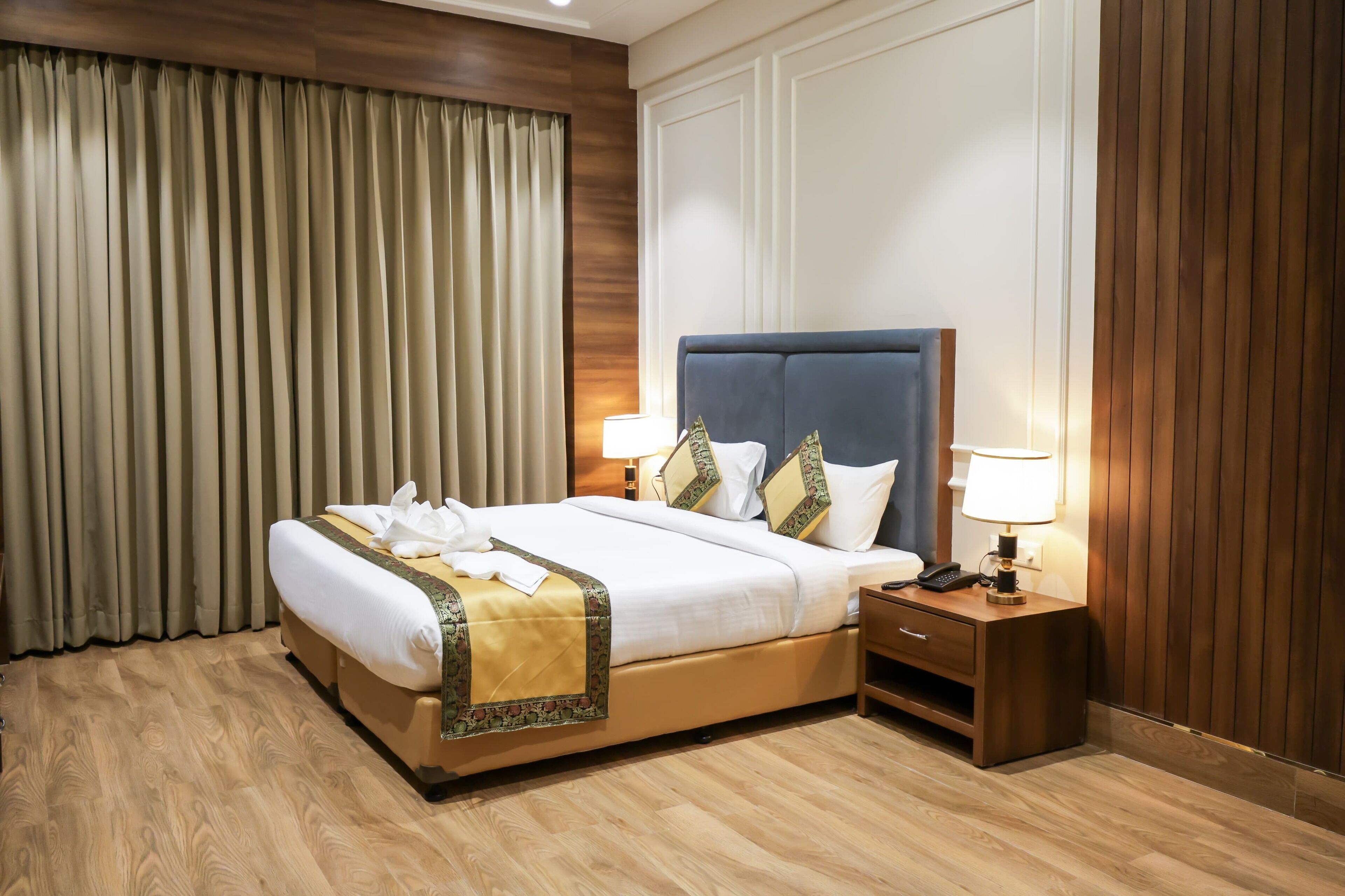 A hotel room with a large bed, wooden flooring, a nightstand with a lamp, and a window with curtains.