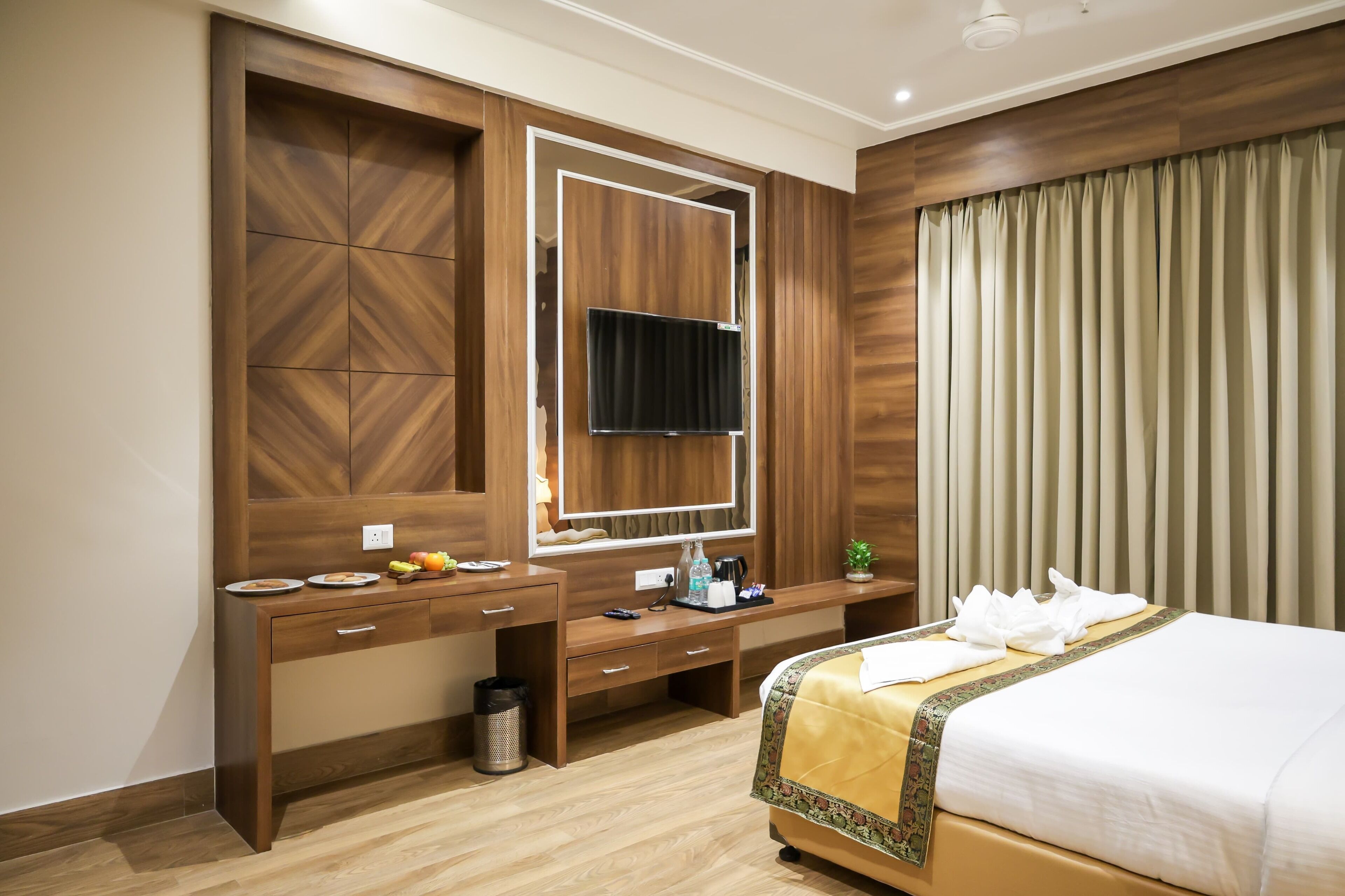 Habitación de hotel con un escritorio de madera, una cama con colcha amarilla, un televisor montado en la pared y un amplio ventanal con cortinas.