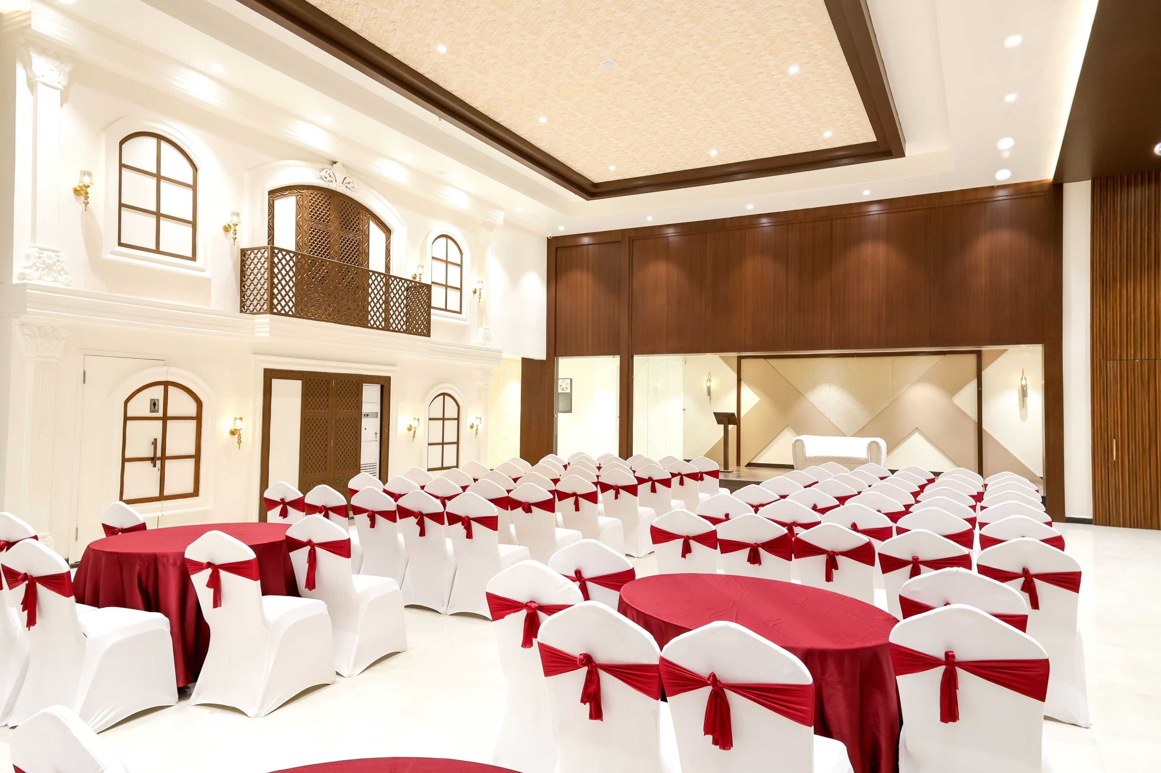 Un salón de conferencias amplio con filas de sillas blancas y manteles rojos, techos altos y un balcón con celosía.