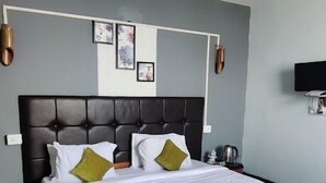 A neatly made bed with a wooden frame, a headboard, and a green throw blanket. There is a bedside table with a lamp and a small decorative item. The room has light blue walls and a wooden floor. - Hotel Valley View by Ravongla Star (Gyalshing)